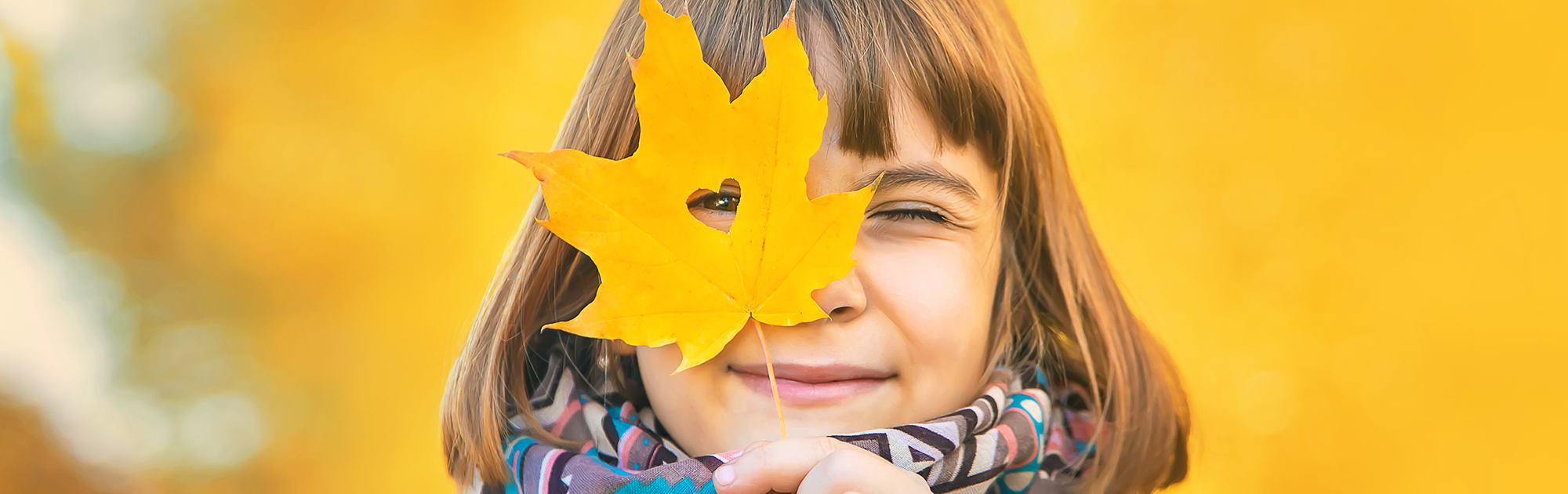 Kind hält ein gelbes Herbstblatt mit Herzform-Ausschnitt vor das Gesicht, herbstlicher Hintergrund.