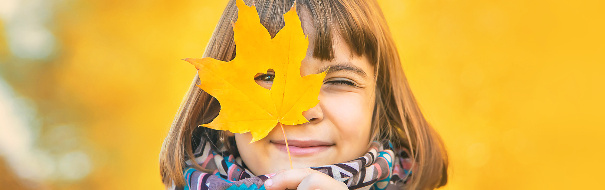 Kind hält ein gelbes Herbstblatt mit Herzform-Ausschnitt vor das Gesicht, herbstlicher Hintergrund.