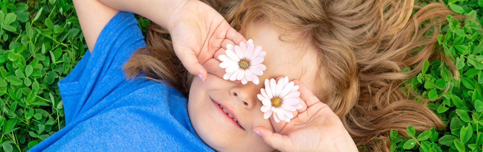 Kind liegt im Gras und hält zwei Gänseblümchen vor die Augen