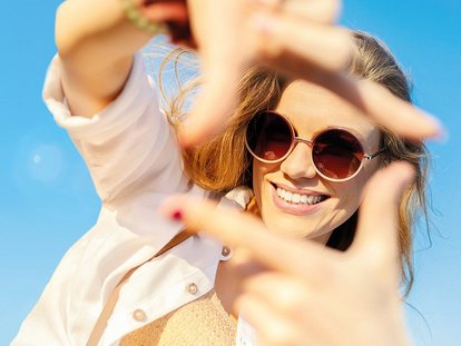 Frau mit Sonnenbrille formt Handrahmen vor sonnigem Himmel
