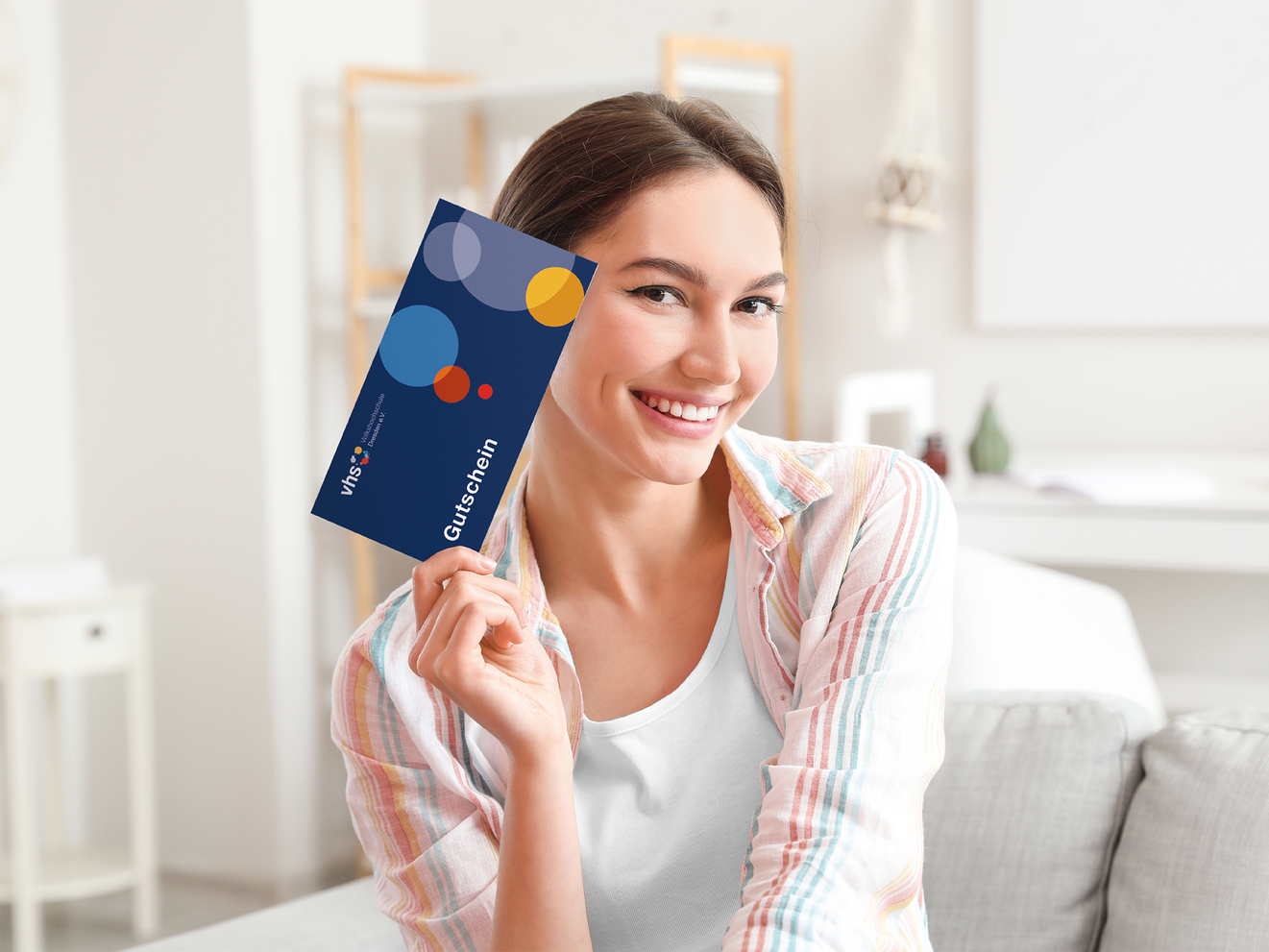 Junge Frau sitzt auf einem Sofa und hält lächelnd einen blauen Gutschein der Volkshochschule Dresden in der Hand